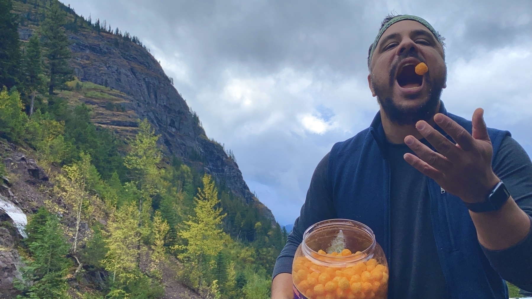 Me in Glacier National Park eating cheeseballs from a big jar with mountains behind me. 