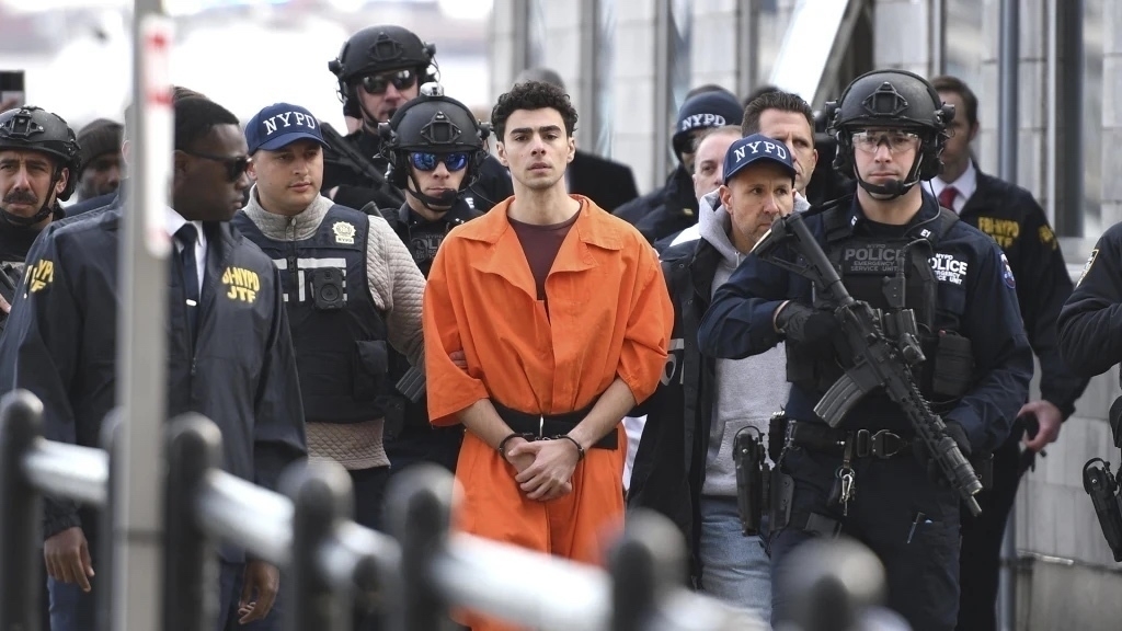 Man in orange jumpsuit handcuffed with armed militarized police around him walking. 