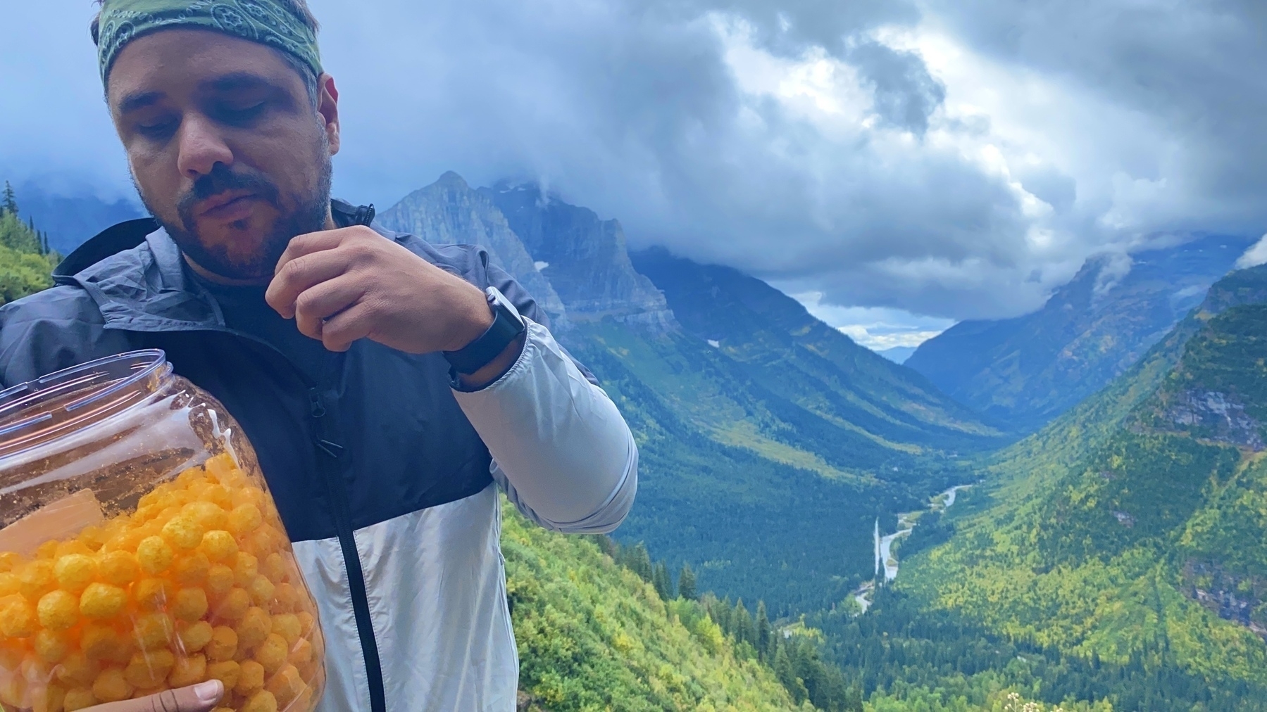 Me in Glacier National Park with a big jar of cheeseballs with mountains behind me. 