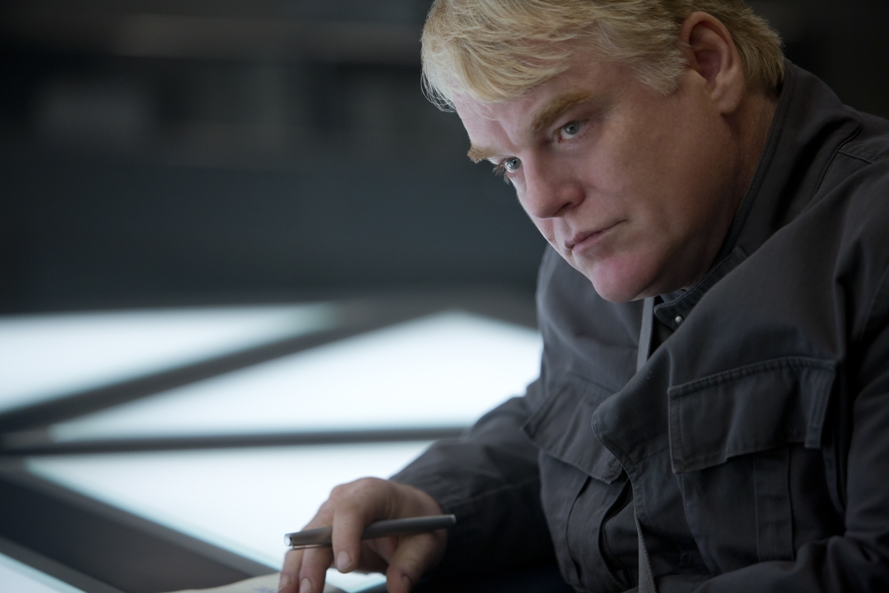 Plutarch from the movie The Hunger Games wearing a dark grey jacket with a pen in his hand. 