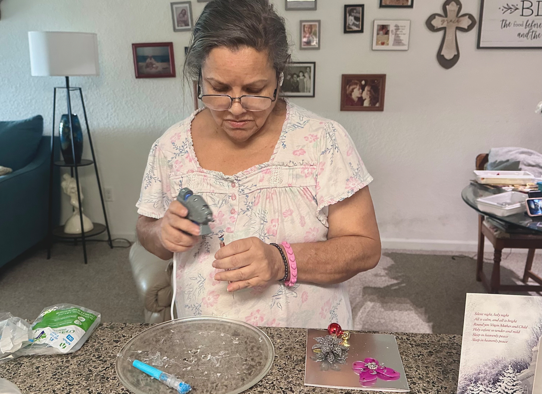 Mom by the counter wearing glasses and AirPods. She has a hot glue gun and other craft tools near a Christmas card that she's making. 