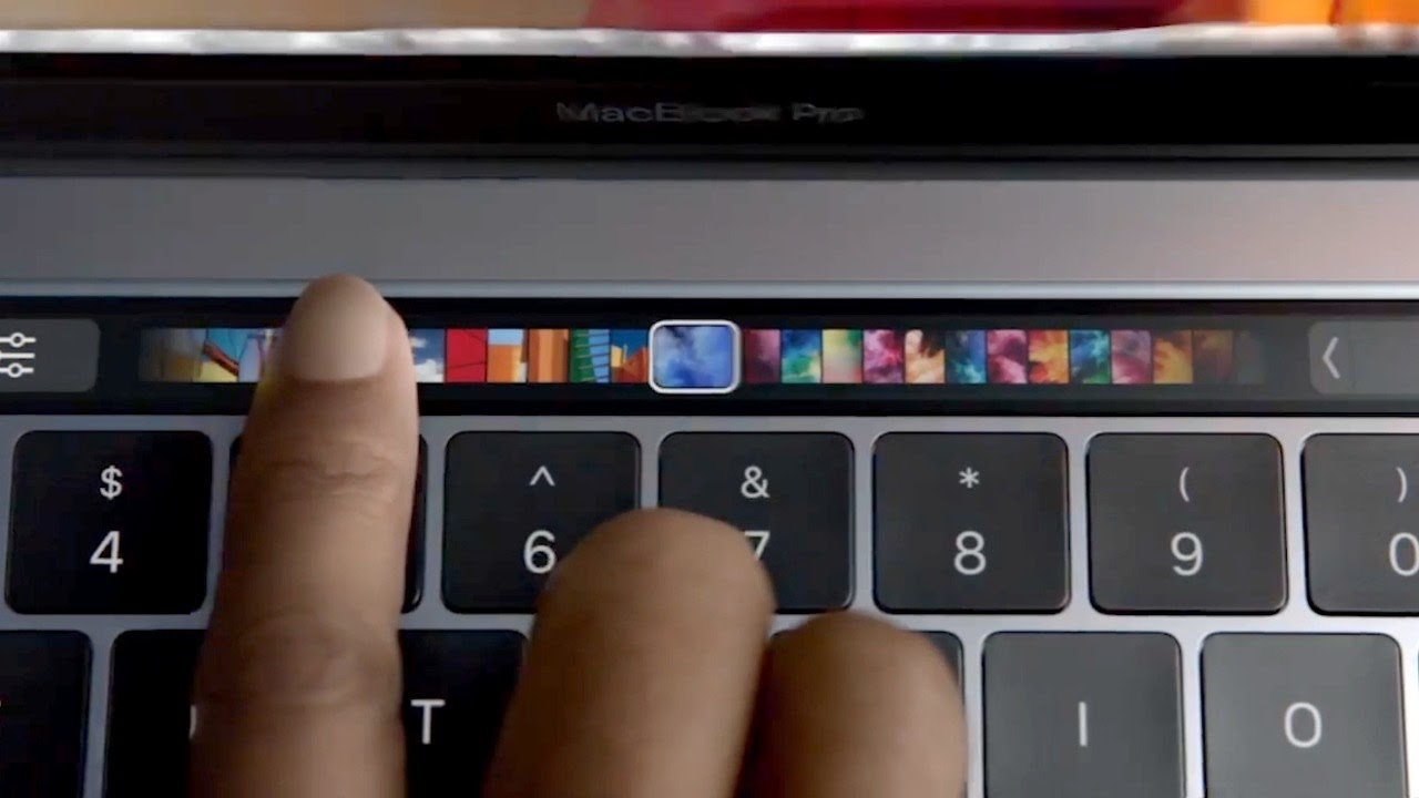 Image of a MacBook Pro keyboard with a hand touching the Touch Bar, displaying various colorful icons and images.