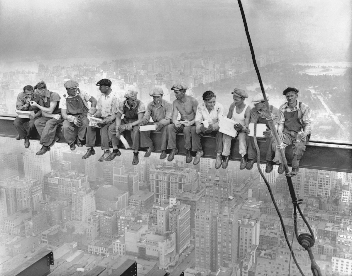 Black and white photo of construction workers sitting on a beam high in the air eating lunch. 