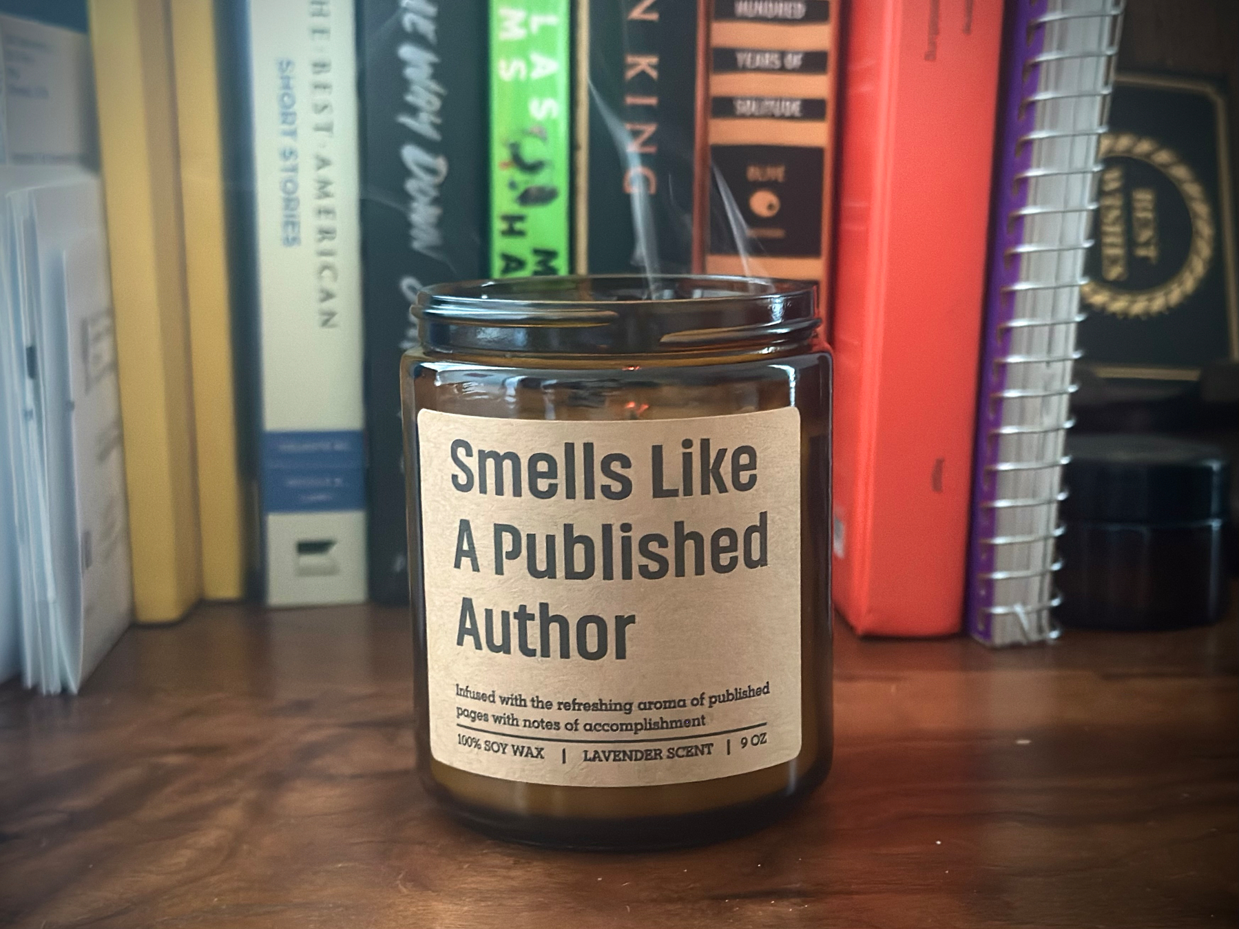 Glass candle, dark tinted. It reads: "Smells like a published author." There's a bit of smoke coming from the top of the candle jar. The jar rests on a mahogany wood surface (my desk). ￼In the background are books, notebooks, and envelopes lined up. 