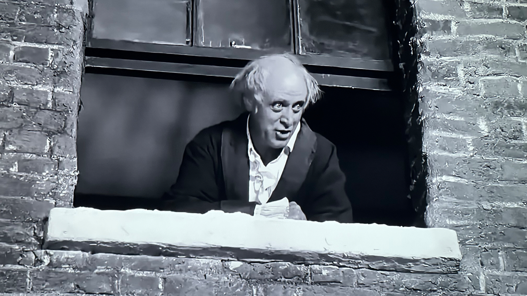 Old man, bald. Wearing a black jacket and white button down pajamas. He's looking out the window excitedly. Black and white photo. 