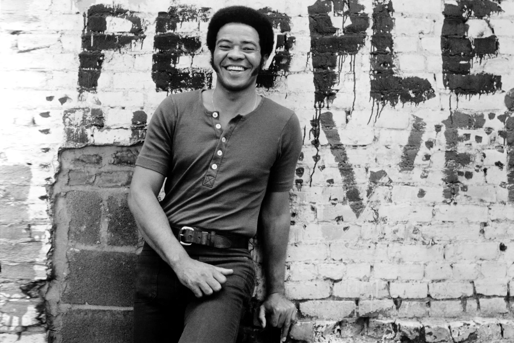 Black and white photo of Bill Withers. He has a medium length haircut. Wearing a Henley t-shirt and jeans. He's smiling. He's leaning on a brick wall behind him. The word "people" is spray painted onto the wall. 