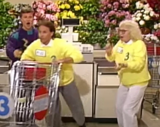 Photo: three people, two contestants in yellow sweaters and one game show host. They're in a supermarket. Flowers in the background. One contestant is about to sprint with their cart, and the other is cheering them on. 