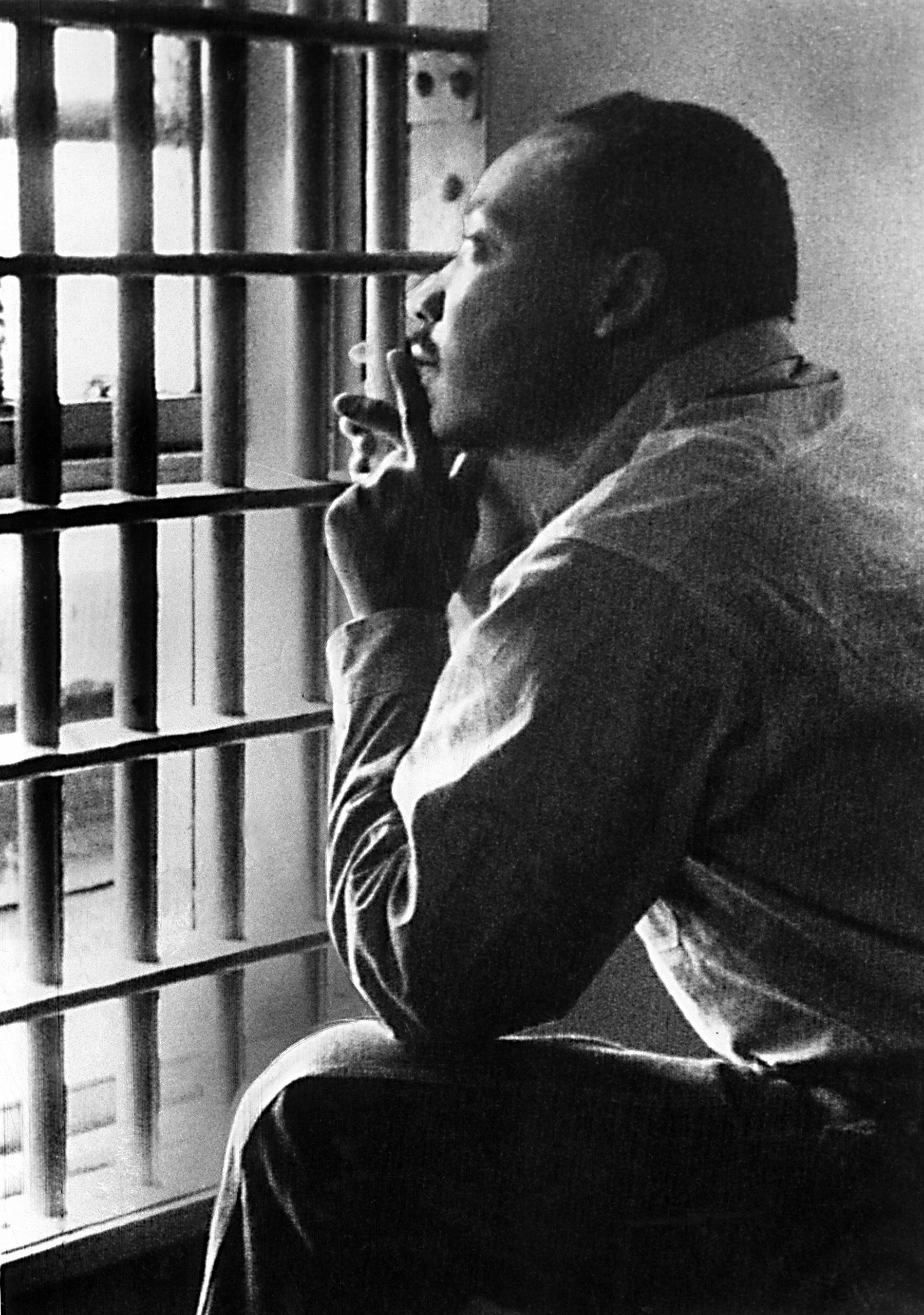 Dr King sitting in a jail cell looking out past the prison bars. He might be smoking a cigarette(?) the photo is in black and white. 
