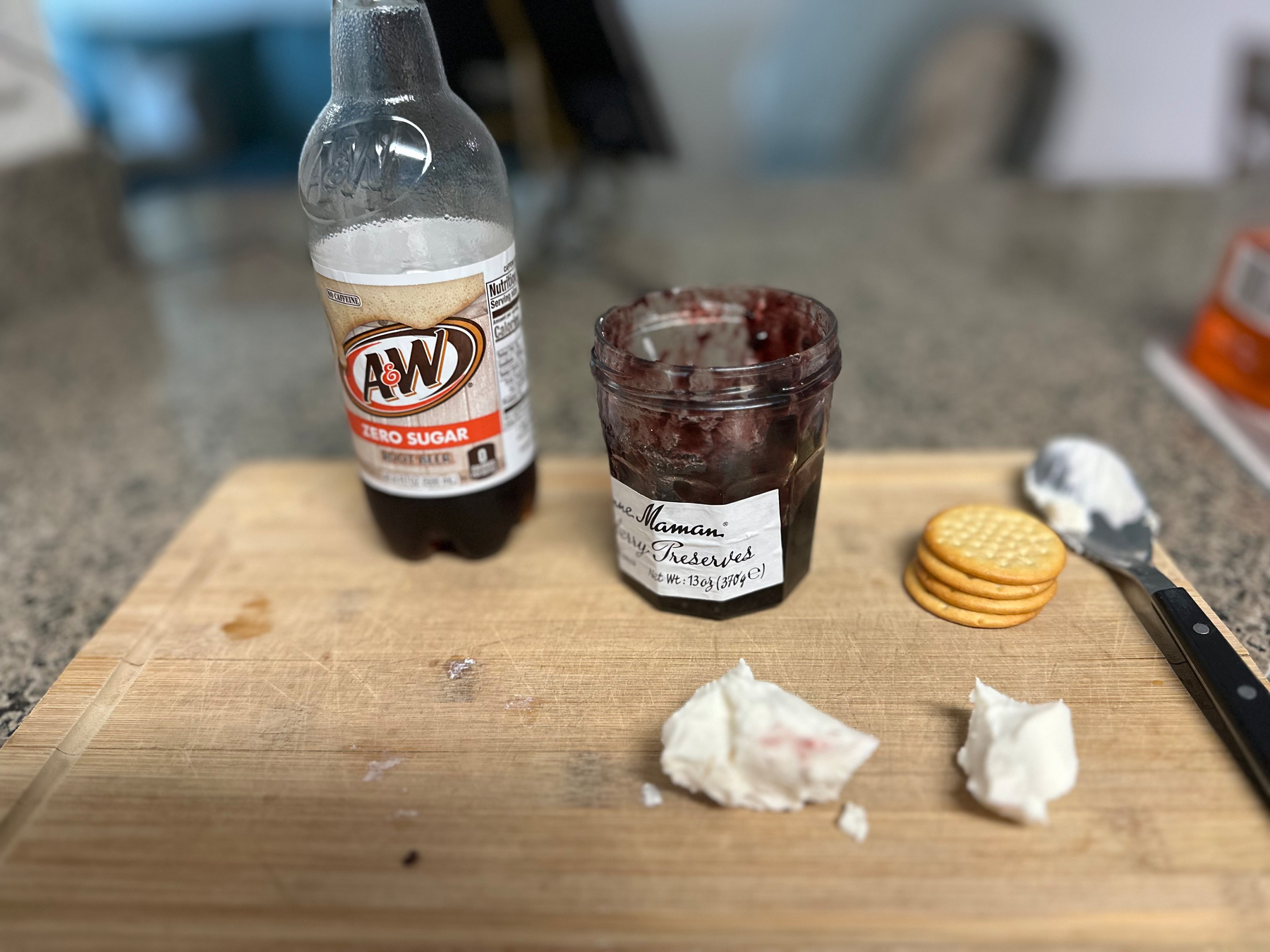 A jar of jam, a pile of crackers, two heaps of goat cheese, and a zero sugar root beer, rest on a wooden cutting board. ￼