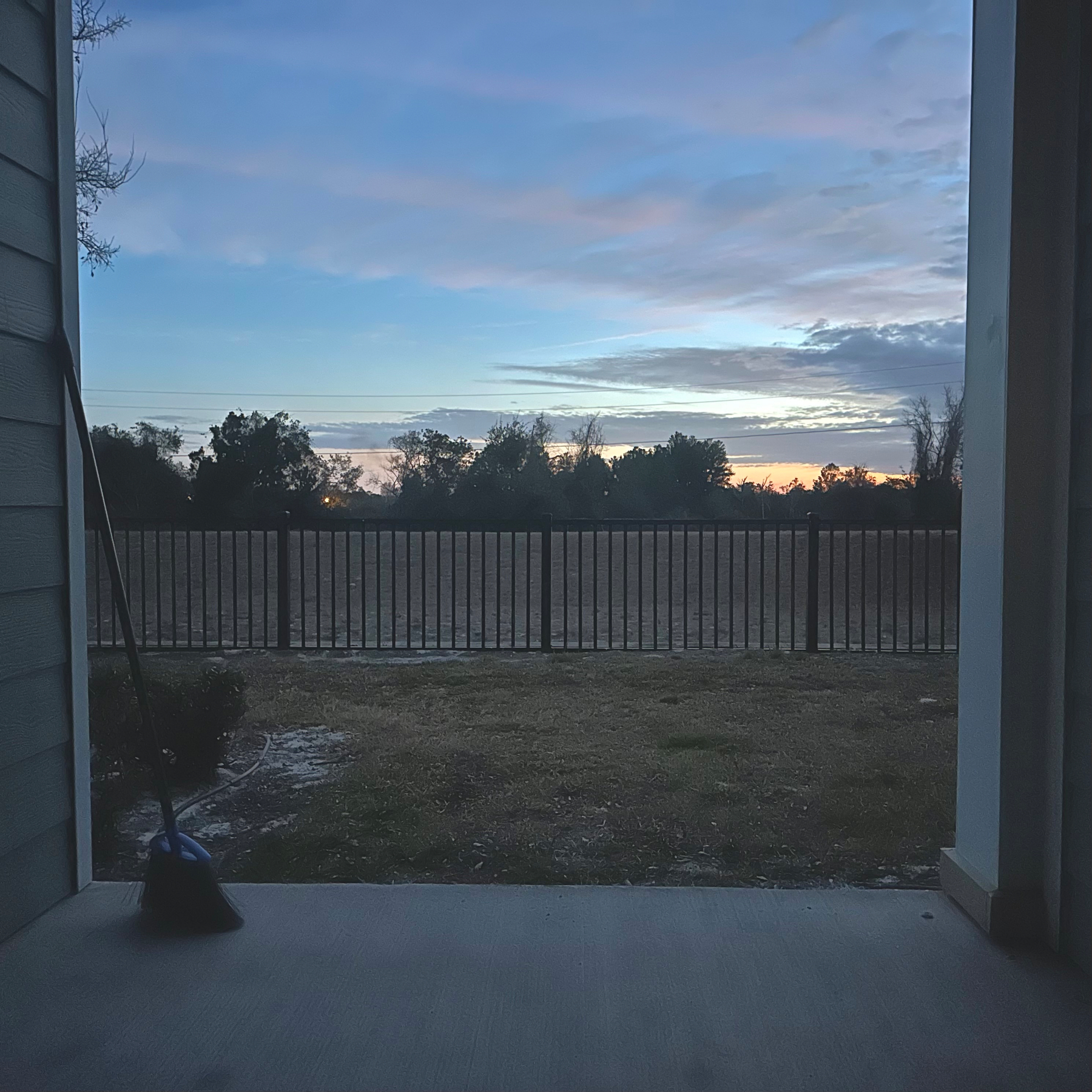 First floor patio out looking a blue and pink sky with trees lining the background. The mid ground has a black fence. There's a broom leaning against the left side of the patio wall. 