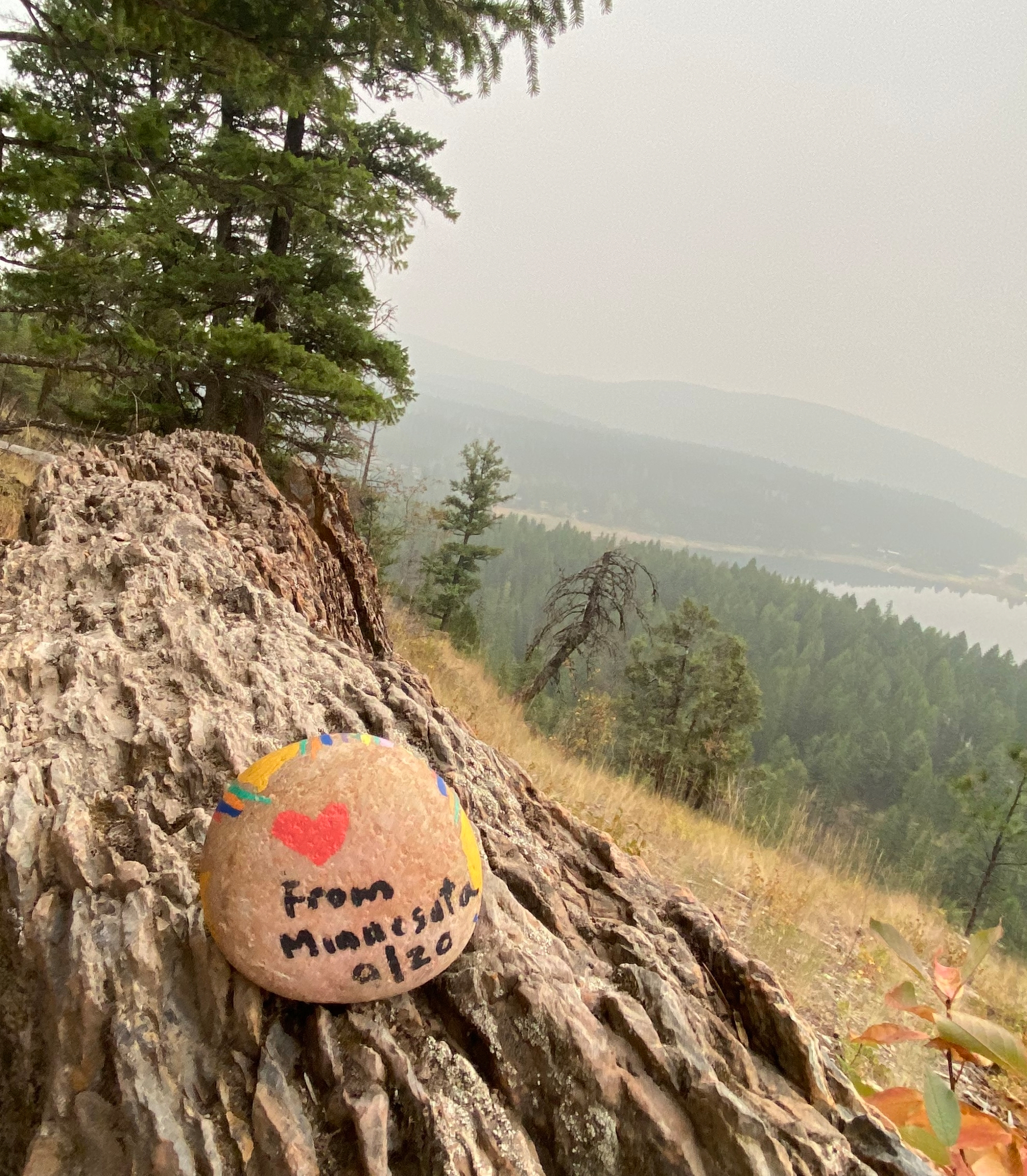 A small smooth rock sits on a rocky floor with mountains and pine trees in the background. The smooth rock have a red heart painted and the message "from Minnesota 9/20"
